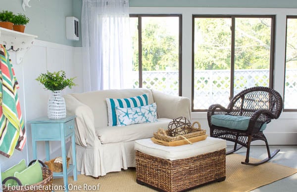 white slipcovered sofa, seagrass ottoman, brown wicker rocking chair, blue and white wall board and batten, striped pool towel, outdoor tan rug 